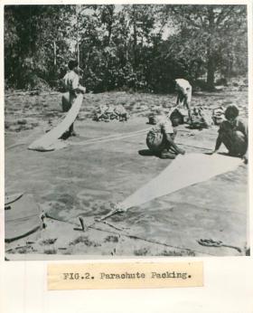 Men from No. 1 Indian Air Company squat on the floor outdoors and pack parachutes.