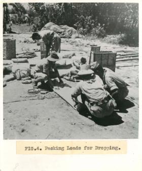 Men from No.1 Indian Air Company squat on teh floor outdoors and pack loads for dropping.