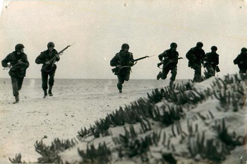 Seven men from 1st Airborne Division run along sandy terrain with guns.