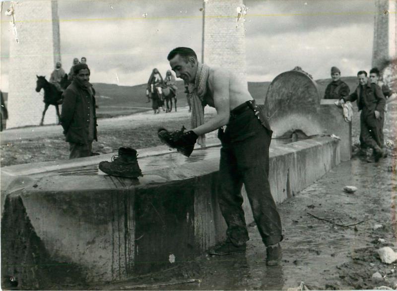 Sergeant Tucker washes his boots in a water trough. Other soldiers and locals look on.  